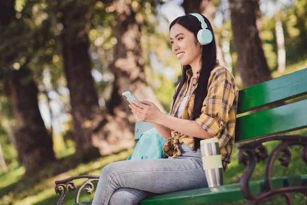 Foto de la encantadora mujer joven vestida camisa a cuadros sentado banco escuchando reproductor de música sonriendo fuera de la naturaleza del campo —  Fotos de Stock