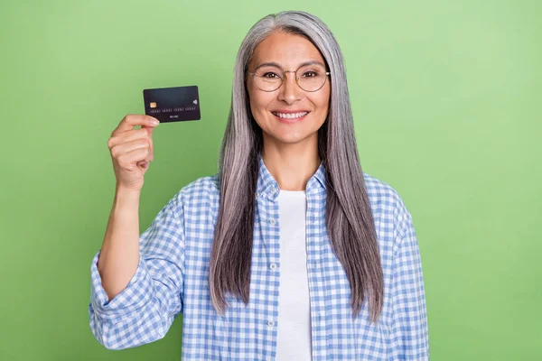 Foto de otimista mais velho penteado cinza senhora mostrar cartão desgaste óculos camisa xadrez isolado no fundo de cor verde — Fotografia de Stock