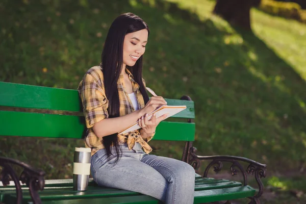 Fotografie okouzlující sladká mladá žena oblečená kostkovaná košile sedí lavice psaní plánovač dělat seznam usmívá mimo přírodu — Stock fotografie