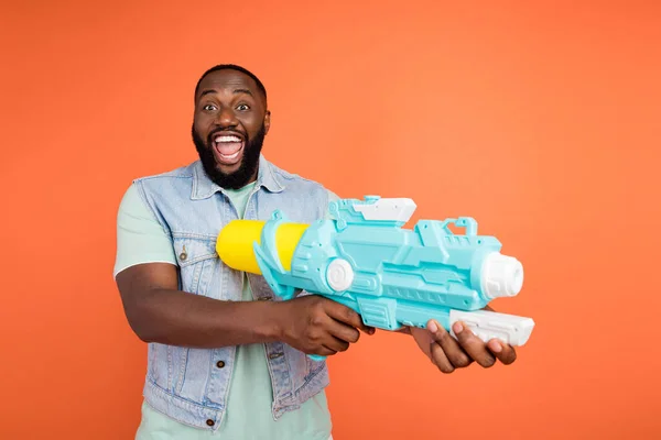 Fotografie funky afro chlap nosit džínovou vestu se zábavnou vodní pistole izolované oranžové barvy pozadí — Stock fotografie