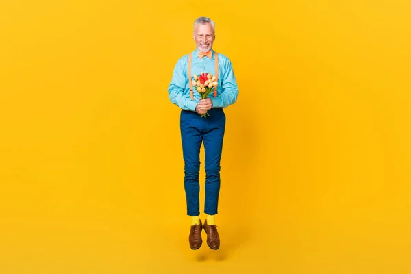Foto de alegre chico lindo pensionista vestido camiseta verde azulado bowtie saltar alto celebración flores aislado color amarillo fondo —  Fotos de Stock