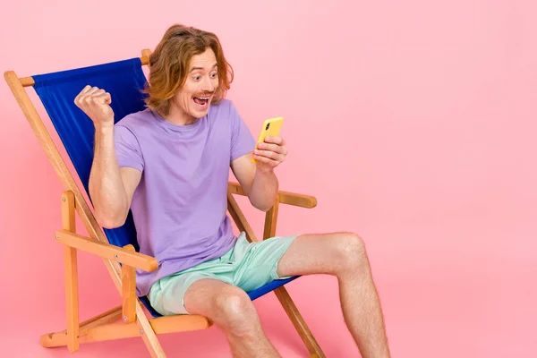 Retrato de chico alegre guapo de moda sentado en silla usando dispositivo celebrando post smm como aislado sobre fondo de color pastel rosa —  Fotos de Stock