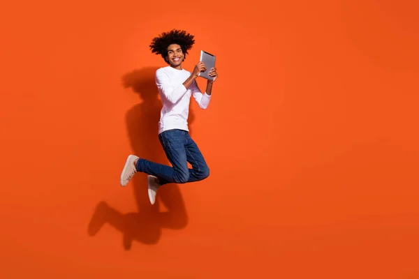 Foto de chico dulce lindo usar camisa blanca saltar alta mecanografía dispositivo moderno espacio vacío aislado color naranja fondo — Foto de Stock