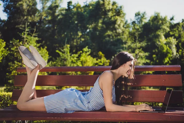 Profil side foto af munter positiv dame liggende bænk udendørs i parken skrive e-mail til ven studerer online på netbook - Stock-foto