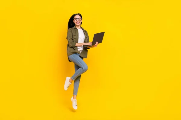 Foto de comprimento total de inteligente jovem senhora salto tipo laptop desgaste eyewear camisa jeans tênis isolado no fundo amarelo — Fotografia de Stock