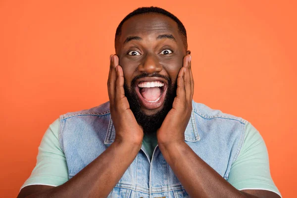 Foto de chico afro lindo impactado usar denim chaleco brazos mejillas boca abierta aislado color naranja fondo — Foto de Stock