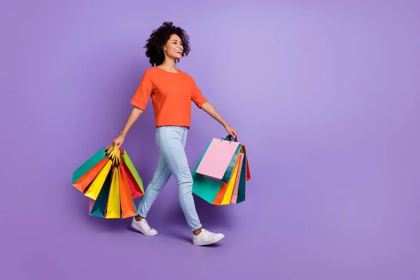 Volledige foto van funky millennial brunette dame gaan met tassen dragen t-shirt jeans sneakers geïsoleerd op violette achtergrond — Stockfoto