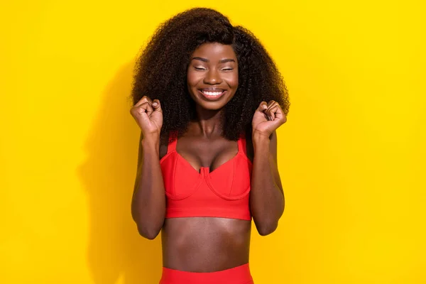Fénykép a szerencsés vicces afro lány viselni piros szexi ruhát emelkedő ököl csukott szemmel mosolygó elszigetelt sárga szín háttér — Stock Fotó