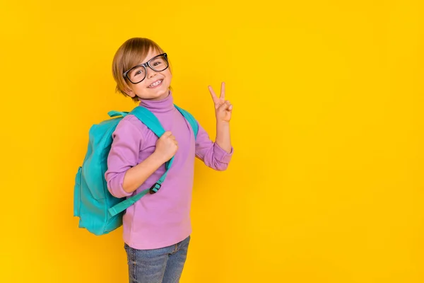 Фото солодкого маленького хлопчика, що показує v-знак, привітається зі своїми новими шкільними друзями ізольовано на жовтому тлі — стокове фото