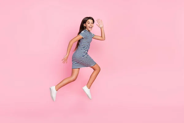 Фото милой дружелюбной девушки носить полосатые платья прыжки высокой бег размахивая рукой изолированный розовый цвет фона — стоковое фото