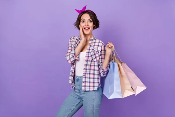 Fénykép lenyűgözött vicces hölgy öltözött kockás ing kar arc gazdaság bevásárlótáskák elszigetelt lila színű háttér — Stock Fotó
