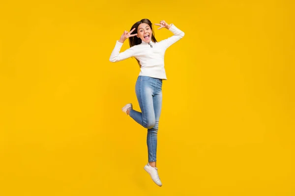 Full Size Foto von süßen brünetten kleinen Mädchen springen Show V-Zeichen tragen Pullover Jeans Schuhe isoliert auf gelbem Hintergrund — Stockfoto