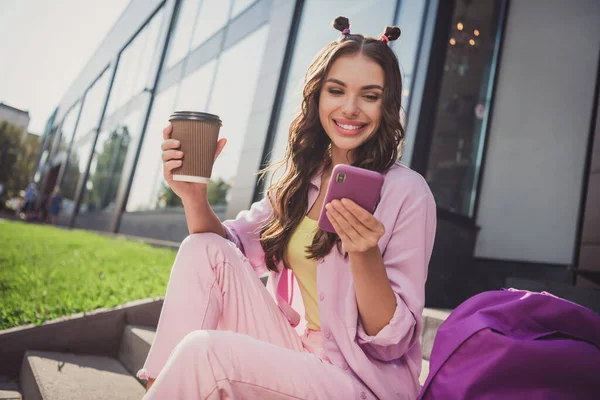 Ritratto di attraente allegra ragazza dai capelli ondulati bere bevanda trascorrere il giorno dopo l'università utilizzando dispositivo all'aperto all'aria aperta — Foto Stock