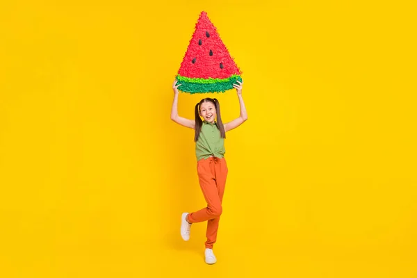 Фото очаровательной девочки-подростка, одетой в зеленый топ, поднимающейся большой кусок арбуза, идущей улыбаясь изолированным желтым цветом фона — стоковое фото