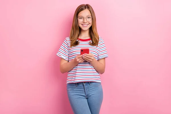 Photo of optimistic small brown hairdo girl hold telephone wear eyewear red t-shirt jeans isolated on pink color background — Stock Photo, Image