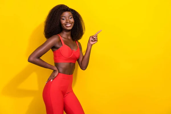 Fotografie sladké hezké afro dívka nosit červené sexy oblečení ukazující prstem prázdný prostor s úsměvem izolované žluté barvy pozadí — Stock fotografie