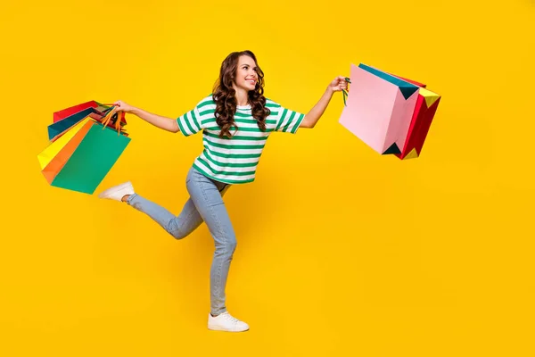 Volledige lichaamsprofiel foto van mooie brunette jonge dame run met tassen dragen t-shirt jeans schoenen geïsoleerd op gele achtergrond — Stockfoto