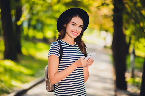 Foto de bonito encantador jovem mulher vestida roupa listrada cap mochila digitando dispositivo moderno andando sorrindo fora da paisagem — Fotografia de Stock