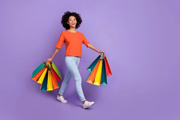 Full size foto van schattige jonge brunette dame gaan met tassen dragen t-shirt jeans schoenen geïsoleerd op violette achtergrond — Stockfoto