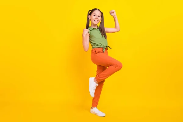 Foto de dulce estudiante emocionada niña usar camisa verde sonriendo puños levantándose caminando aislado color amarillo fondo — Foto de Stock