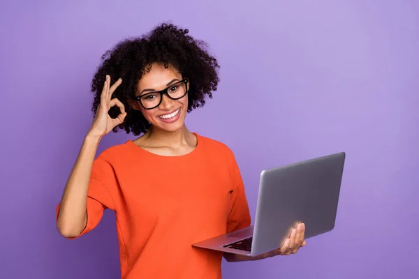 Photo de jeune femme brune impressionnée tenir ordinateur portable okey porter lunettes t-shirt orange isolé sur fond violet — Photo
