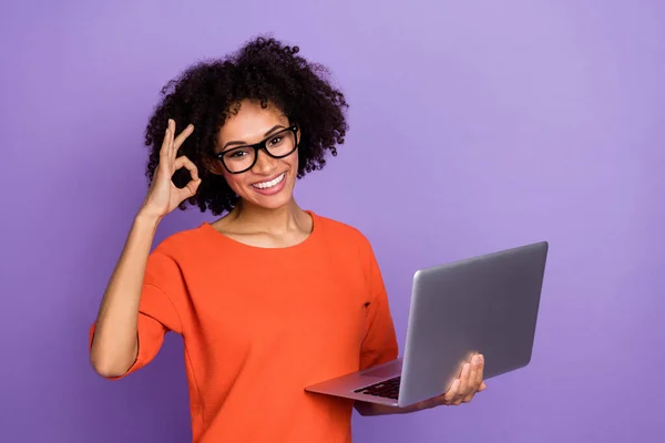 Foto de la joven morena impresionada sujetar portátil okey usar gafas naranja camiseta aislada sobre fondo púrpura —  Fotos de Stock