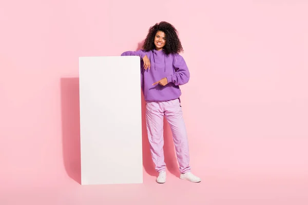 漂亮闪亮的非洲女孩穿着紫色运动衫，手指指向白色广告牌，微笑着孤立的粉色背景的照片 — 图库照片