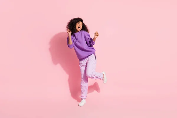 Foto de la afortunada chica afro encantadora usar sudadera violeta bailando buscando espacio vacío levantando puños sonriendo aislado color rosa fondo —  Fotos de Stock