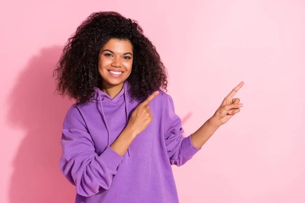 Фото очаровательной милой афро-девушки носить фиолетовую толстовку указывая двумя пальцами пустое пространство улыбаясь изолированный розовый цвет фона — стоковое фото