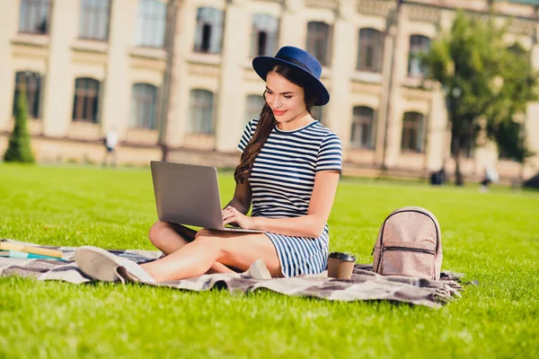 Zdjęcie uroczej młodej kobiety ubranej w paski czapka plecak siedzi trawa odrabiając pracę domową nowoczesne urządzenie poza krajobrazem — Zdjęcie stockowe