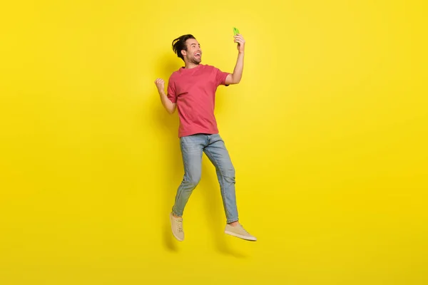 印象的なひげミレニアル男ジャンプの完全な長さの写真電話を見て黄色の背景に隔離された赤いTシャツジーンズの履物を着用 — ストック写真
