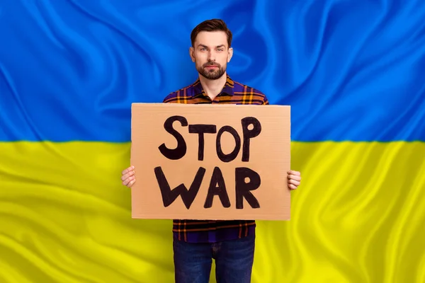 Photo image of young activist guy ask international diplomacy help Ukraine in struggle against russia terrorism repression hold poster yellow blue flag background