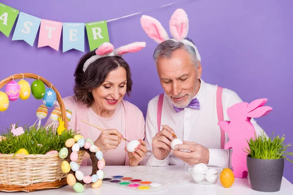 Portrét dvou pozitivních prarodičů malovat barevné vejce připravit událost izolované na fialovém pozadí — Stock fotografie