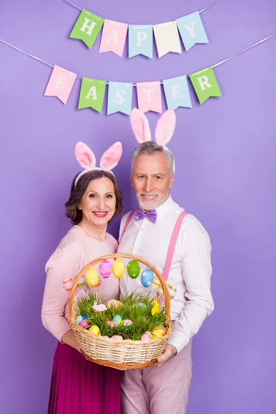 Pionowe zdjęcie dwóch starszych wesołych ludzi trzymać jaj kosz królicze uszy izolowane na fioletowym tle koloru — Zdjęcie stockowe