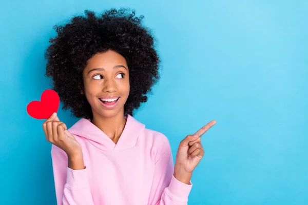 Retrato de chica alegre atractiva sosteniendo tarjeta de corazón demostrando espacio de copia aislado sobre fondo de color azul brillante —  Fotos de Stock