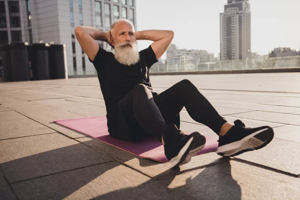 Foto důchodce stařec sedět fitness mat do abs crunches kardio trénink ruce hlava nosit tričko městský město venku — Stock fotografie