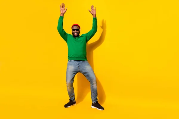 Foto van positieve man dans verhogen handen dragen zonneglas rood muts groen shirt jeans geïsoleerde gele kleur achtergrond — Stockfoto