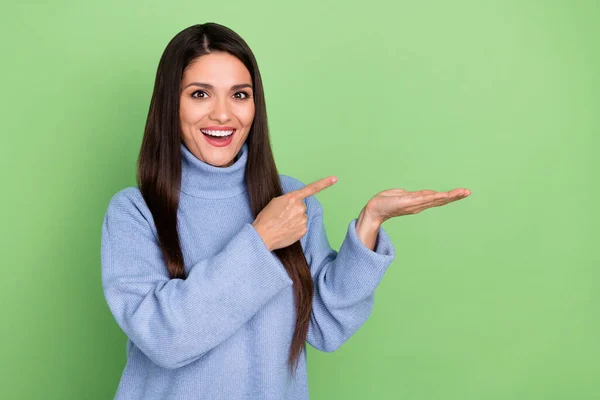 Foto de muito animado jovem mulher usar camisola azul sorrindo apontando dedo segurando braço vazio espaço isolado cor verde fundo — Fotografia de Stock