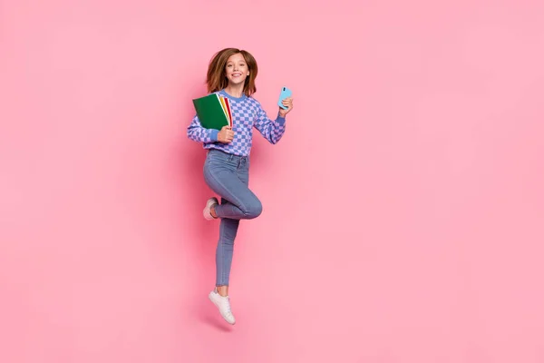 Foto de corpo inteiro de loiro adolescente menina salto segurar telefone livro desgaste camisola jeans sapatos isolados no fundo rosa — Fotografia de Stock