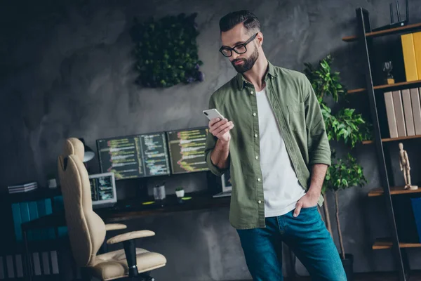 Фотографія зайнятого розумного молодого чоловіка в зеленій сорочці окуляри, що друкують сучасний пристрій в приміщенні робочої станції майстерні — стокове фото