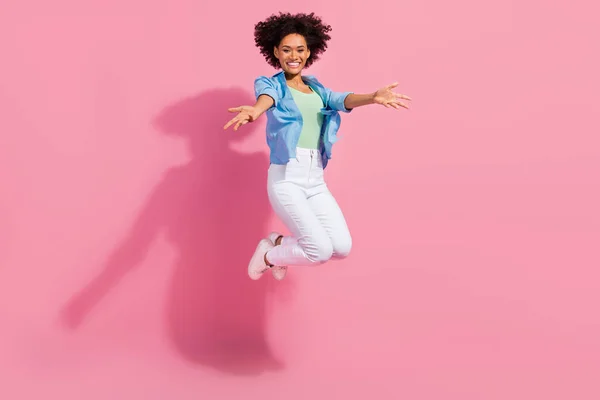 Фото положительной привлекательной леди прыжок растяжения приветственные руки носить джинсовые брюки рубашки кроссовки изолированный розовый цвет фона — стоковое фото