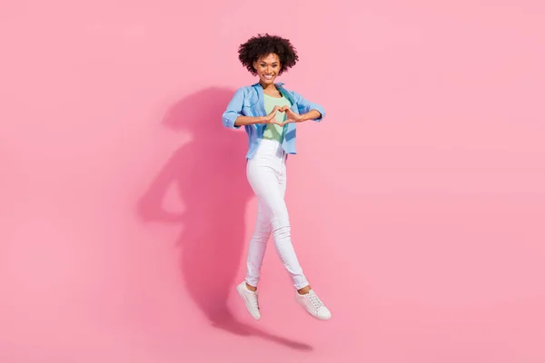 愛情のある女性のジャンプの指の写真は、ハートシンボルは、デニムシャツのズボンのスニーカーを着用ピンクの色の背景を示す — ストック写真