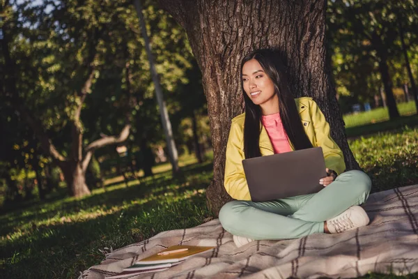 Портрет привлекательной веселой мечтательной девушки, сидящей на обложке с помощью ноутбука, проводящей выходные за просмотром фильмов на открытом воздухе — стоковое фото