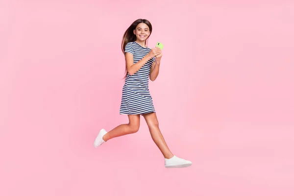 Foto de linda chica adorable vestido a rayas chateando gadget moderno saltar alto aislado color rosa fondo —  Fotos de Stock