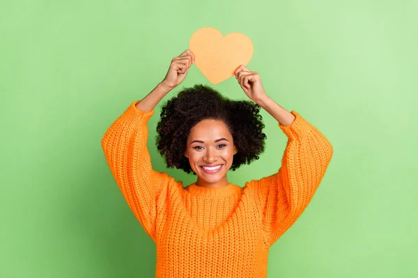 Fénykép az édes fiatal hullámos frizura hölgy tartsa szív viselni narancssárga pulóver elszigetelt zöld színű háttér — Stock Fotó