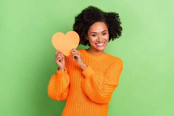 Foto de flirty millennial rizado peinado dama espectáculo corazón desgaste naranja suéter aislado en fondo de color verde —  Fotos de Stock