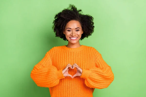 Foto di flirty millennial capelli ricci signora spettacolo cuore usura arancione maglione isolato su sfondo di colore verde — Foto Stock