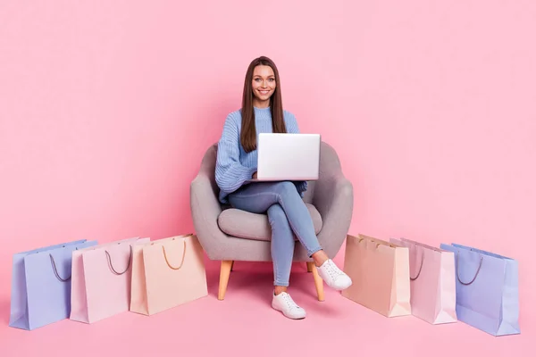 Фотография в полный рост, на которой девушка сидит в кресле с помощью ноутбука, со скидкой в магазине на фоне розового цвета — стоковое фото