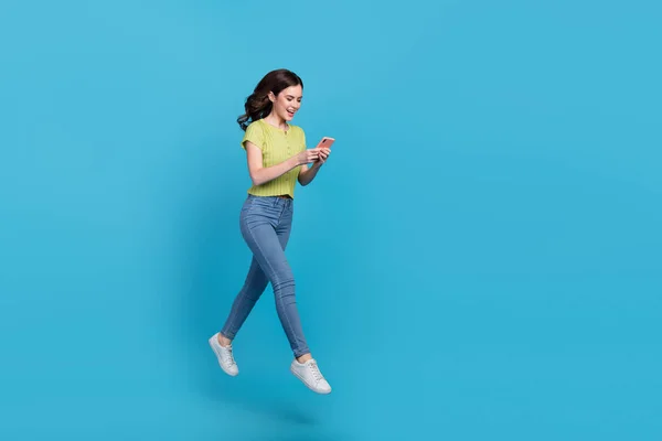 Full längd foto av söta unga brunett dam springa titta telefon slitage t-shirt jeans sneakers isolerad på blå bakgrund — Stockfoto