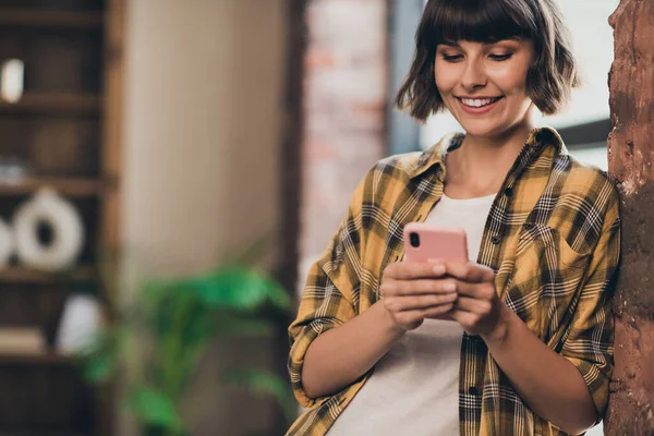 Foto de adorable dulce joven mujer vestida camisa a cuadros mecanografiando gadget moderno en el interior de la estación de trabajo — Foto de Stock